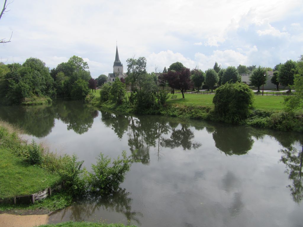 La rivière de l'Eure à Léry, bordée d'arbres, avec le clocher de l'église Saint-Ouen à l'horizon
